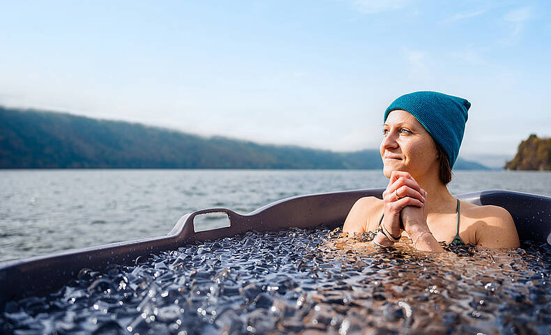Frau sitzt in einer Holzbadewanne voller Eiswasser und genießt Ausblick auf den Millstätter See