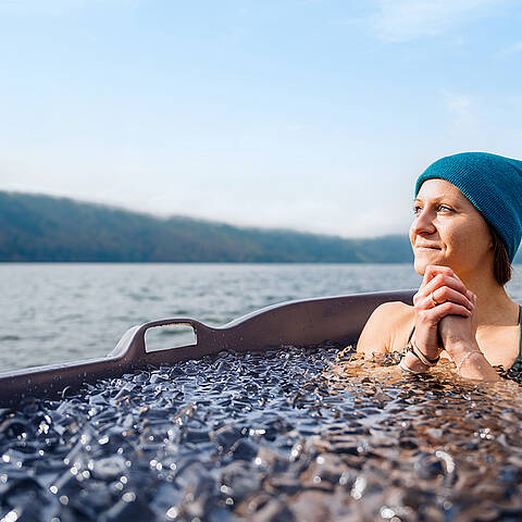 Frau sitzt in einer Holzbadewanne voller Eiswasser und genießt Ausblick auf den Millstätter See