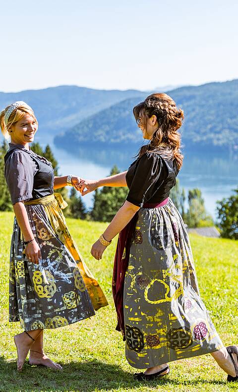 2 Frauen tanzen auf einer Wiese in Tracht mit Blick auf den Millstätter See
