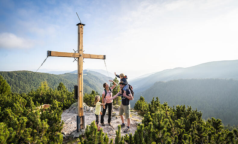Familie mit Kindern bei einer Wanderung in den Nockbergen, die glücklich an einem Gipfelkreuz steht und die Aussicht genießt.